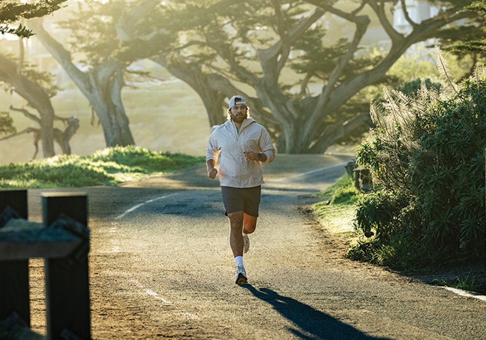 Person running on a paved path through a park-like setting, wearing a smartwatch, with large trees and soft morning light in the background.