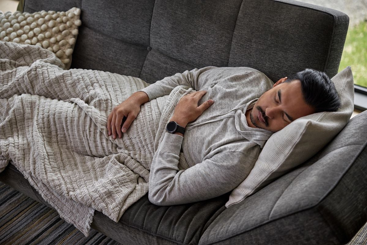 A man in a gray shirt naps on a dark gray couch with a cream colored blanket.