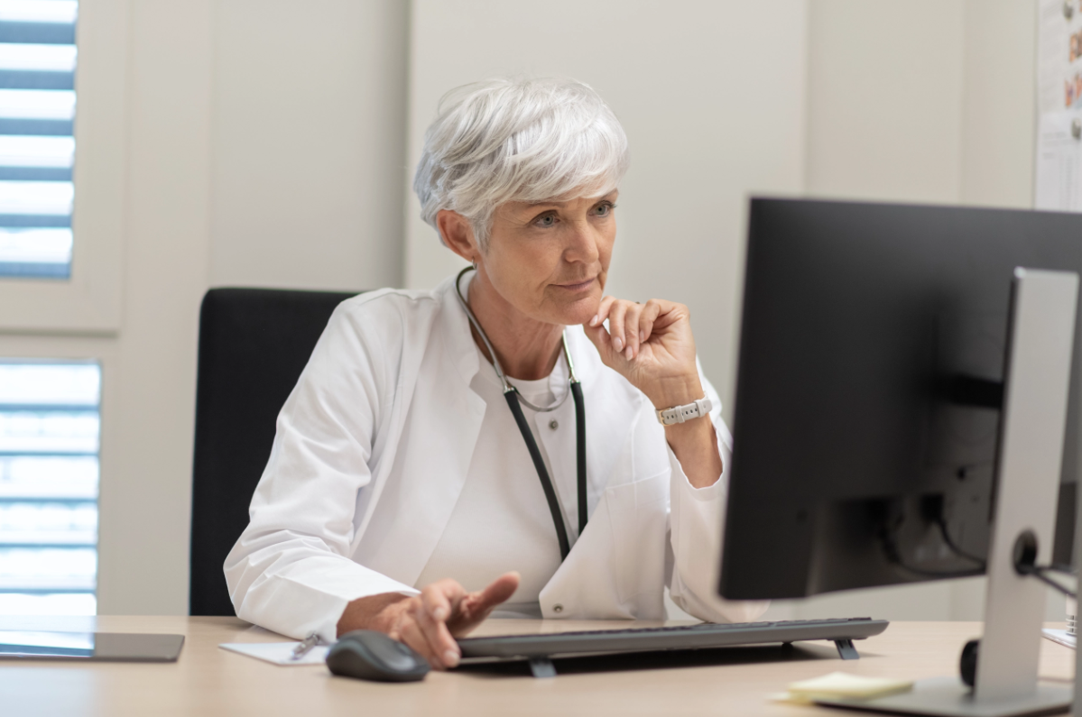 A female doctor sits at a desk in an office and looks intently at a computer monitor.