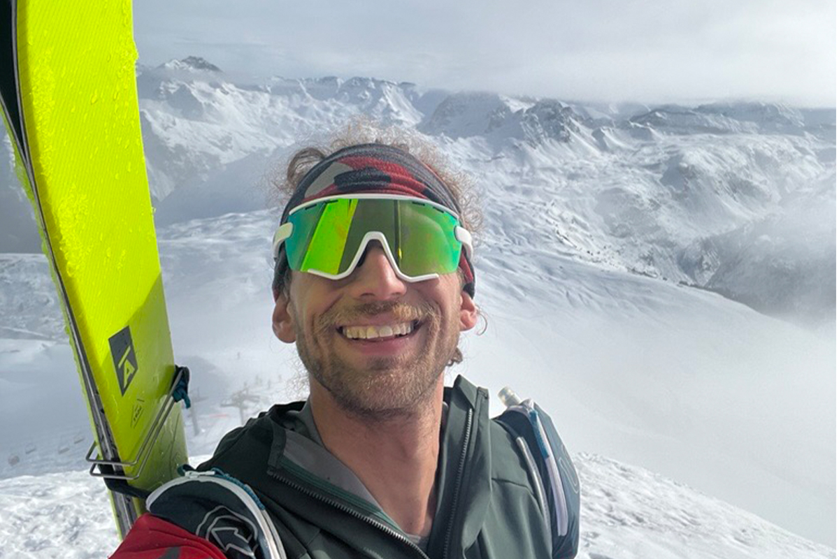 Skier Noah Dines, carrying bright yellow-green skis, takes a selfie in the snow-covered mountains.