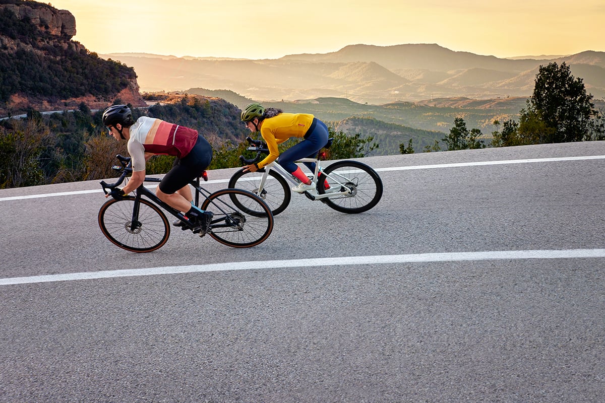 Two cyclists ride bikes on a road with mountains in the background.
