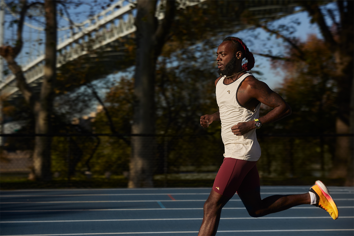 A man in a white tank top and red shorts runs with a bridge in the background.