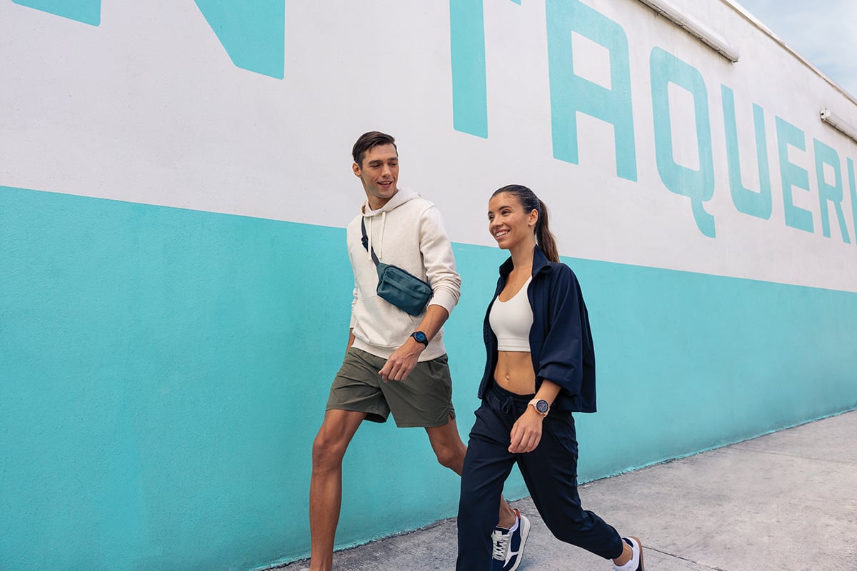 A man in a cream sweatshirt and green shorts walks next to a smiling woman wearing a navy jacket and pants.