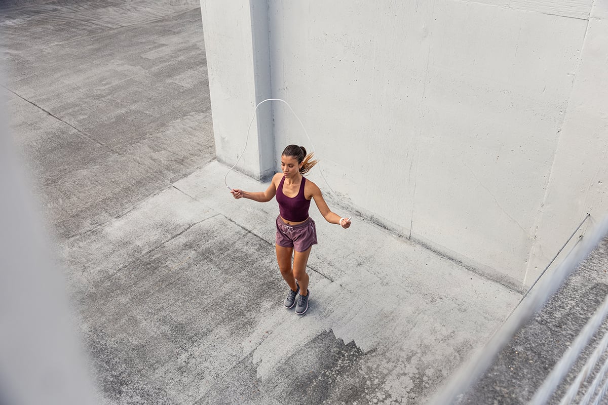A woman wearing a purple tank top and light purple shorts exercises by using a jump rope.
