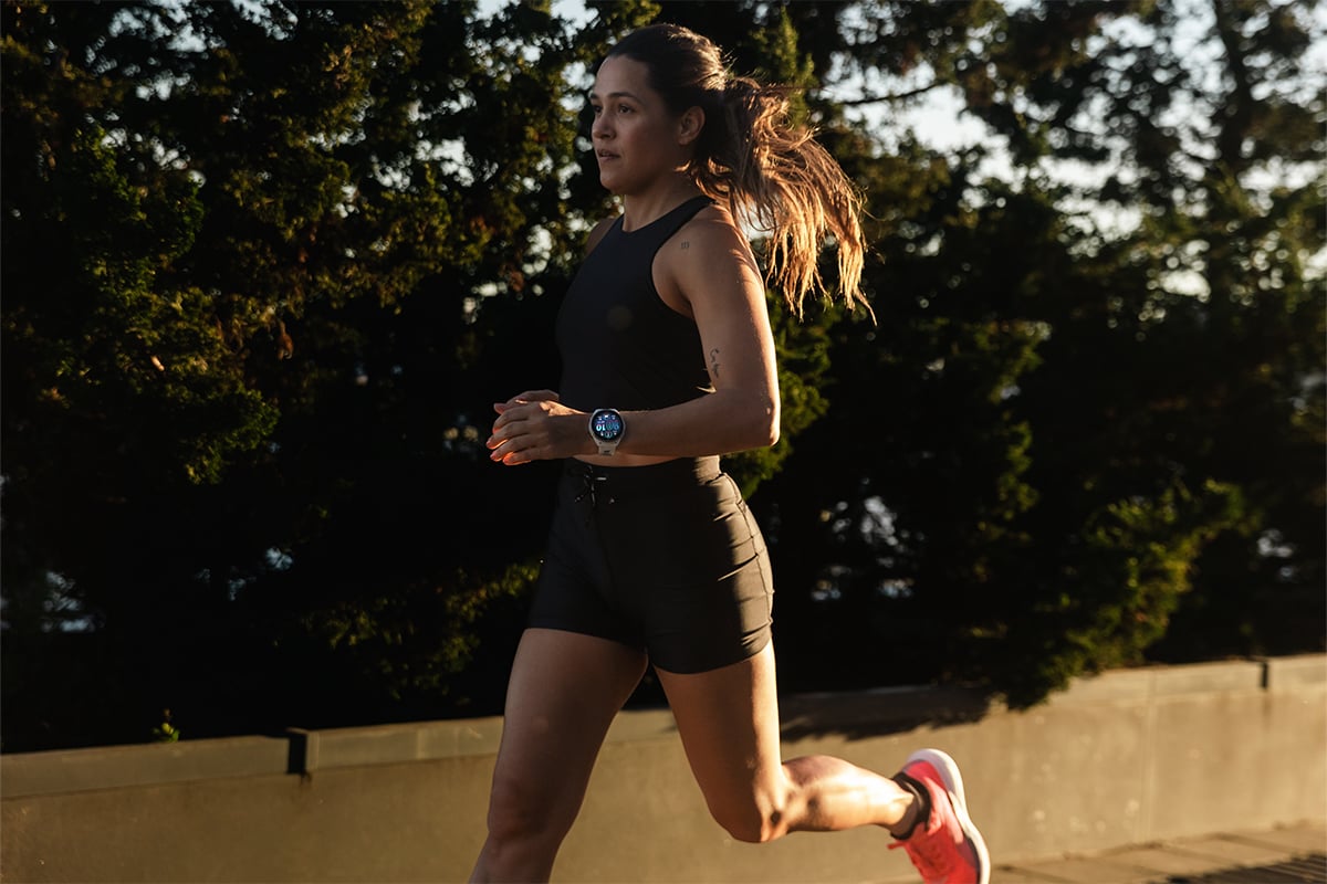 A woman wearing a black tank top and shorts and orange shoes runs with trees in the background.