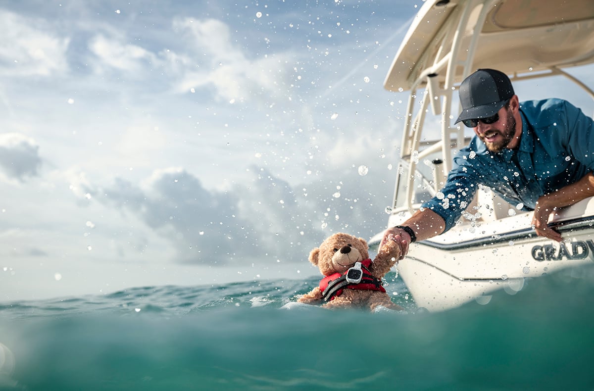 A man reaching into the water from his boat grabbing a teddy bear out of the water. The teddy bear has an MOB tag and life jacket on.
